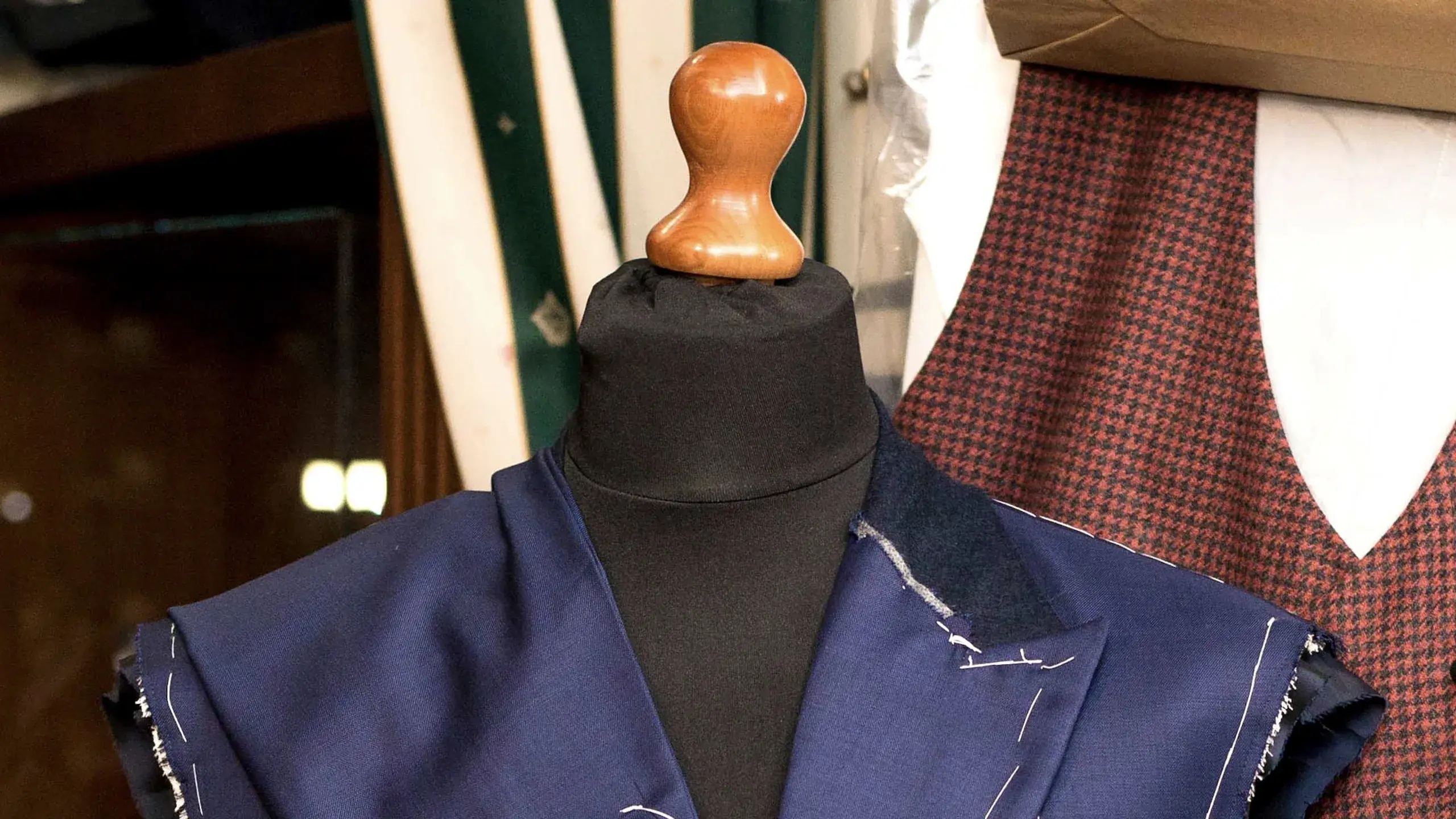 A navy jacket basted in white thread on a tailor's stand, a houndstooth waistcoat hanging behind it