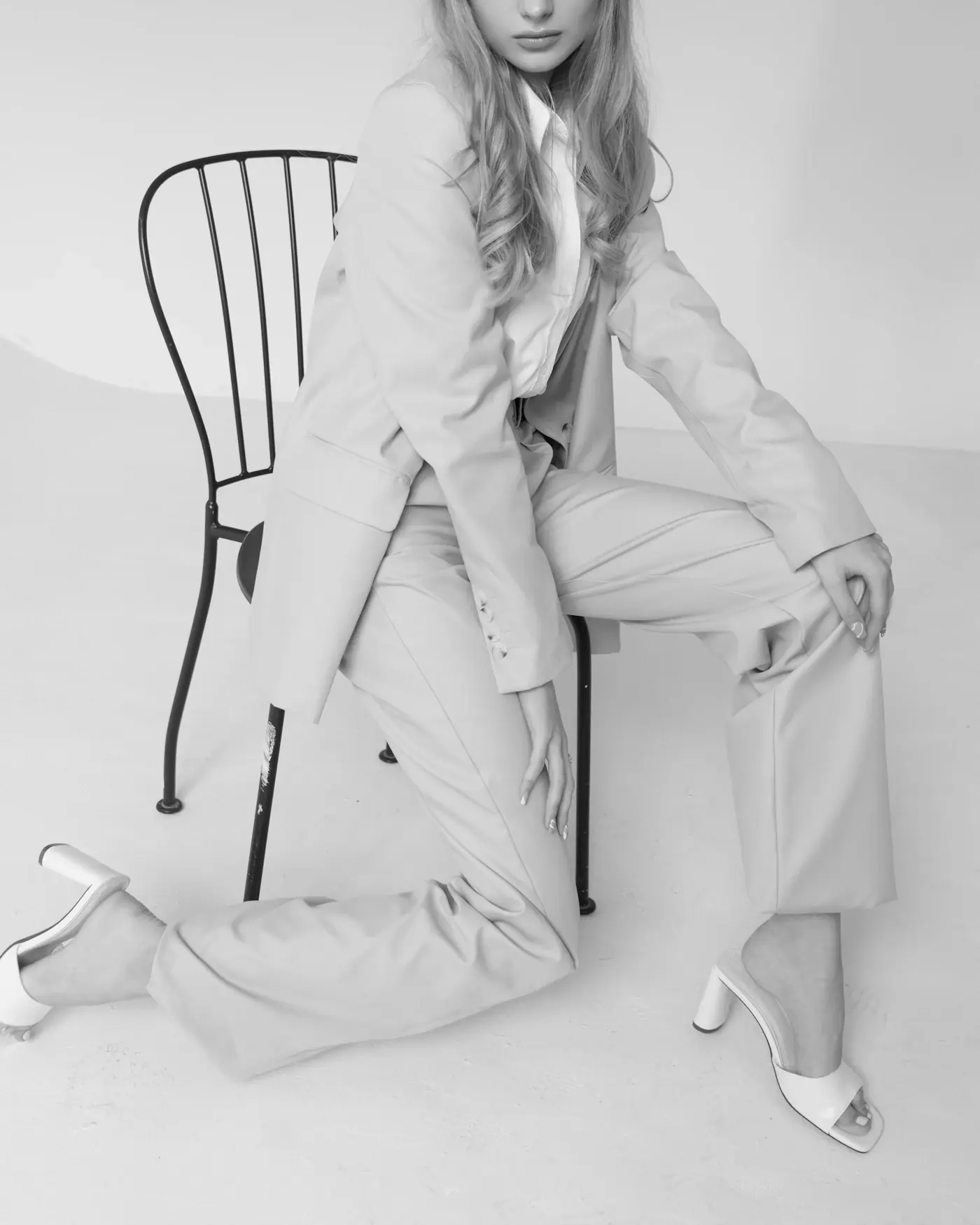 A model in a pale suit seated on a steel chair in a white studio, in black and white