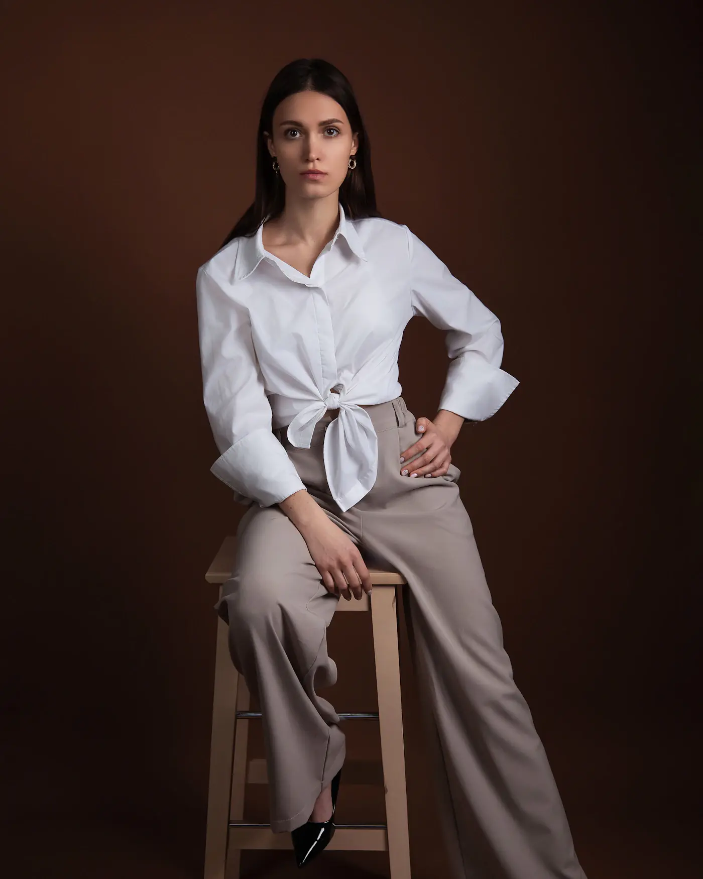 A model seated on a wooden stool in wide stone trousers and a white shirt